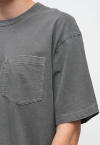 Szürke pamut rövid ujjú póló környakkal és mellzsebbel. Laza szabású, puha tapintású. A szélek enyhén bolyhozottak.