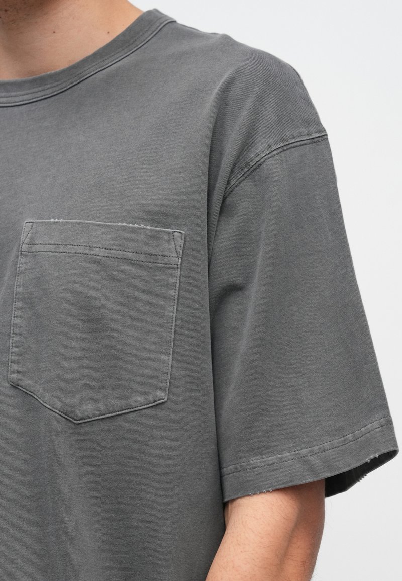 Grå bomulls-T-shirt med rund hals, korta ärmar och en bröstficka. Har en avslappnad passform och mjuk textur. Kanter något fransiga.