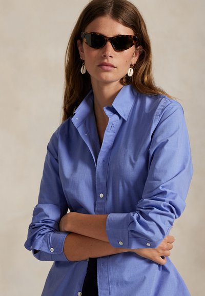 Kvinne med langt brunt hår som har på seg skilpaddemønstrede solbriller, hvite skjelløreringer og en blå knappeskjorte med oppbrettede ermer.