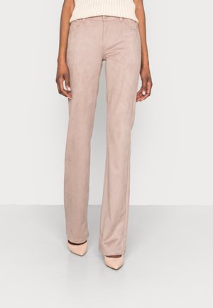 Vrouw die lichtroze rechte broek en naaldhakken met een spitse neus draagt, staand tegen een effen achtergrond.