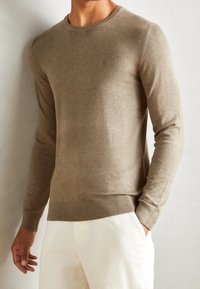 Beżowy sweter robiony na drutach z okrągłym dekoltem, długimi rękawami oraz prążkowanymi mankietami i dołem. Tkanina wydaje się miękka i może mieć subtelną fakturę.