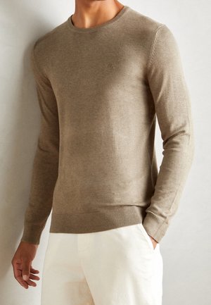 Jersey beige de punto con cuello redondo, mangas largas y puños y dobladillo acanalados. La tela parece suave y puede tener una sutil textura.