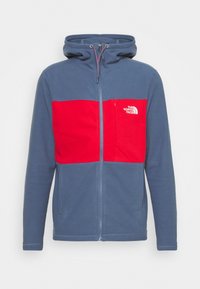 Giacca in pile blu con un pannello toracico e tasche rosse, dotata di zip anteriore e cappuccio. Include un dettaglio del logo sul lato sinistro.