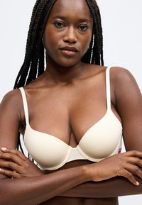 Béžová podšitá podprsenka s nastaviteľnými ramienkami, podporným oblúkom a bezšvovým dizajnom. Textúrovaná tkanina s hladkým povrchom. Minimálne kovové detaily.