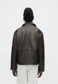 Brūna ādas jaka ar gludu tekstūru, tai ir apkaklveida izgriezums un kastes formas siluets. Uz muguras redzamas šuvju detaļas.