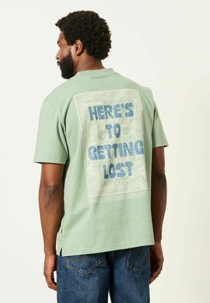 Man met kort krullend haar en baard die een lichtgroen t-shirt draagt met blauwe tekst ""HERE'S TO GETTING LOST"" op de rug.