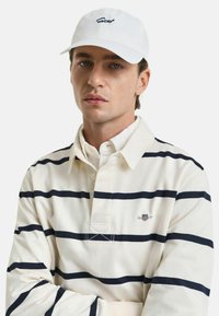 Witte baseballpet met geborduurd logo, gecombineerd met een crèmekleurige longsleeve polo met marineblauwe horizontale strepen en een met knopen sluitende kraag.