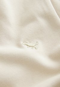 Beež tekstiil pehme, tekstuurse pinnaga, mille keskel on väike heledast niidist tikitud mustriline detail.