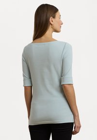 Haut ajusté bleu clair avec des manches 3/4 et des poignets retroussés, présentant un large décolleté et une texture douce et lisse. Vue de dos montrée.