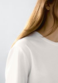 Hvid bomulds t-shirt med rund hals, glat tekstur og korte ærmer, delvist vist med langt lige hår i baggrunden.