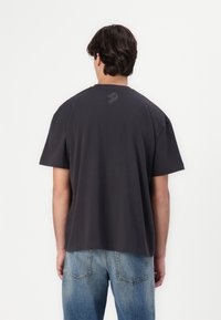Dark gray cotton t-shirt with a relaxed fit, short sleeves, and a small decorative pattern on the upper back. Paired with blue jeans.