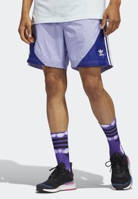 Shorts in rete viola e blu navy con pannelli di colore contrastante e tre strisce. Scarpe sportive nere con una suola rosa, abbinate a calzini viola tie-dye.