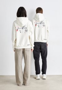 Hoodie crème avec un graphique "MILANO" en noir et des accents de splatter. Associé à un jean large gris clair et noir.