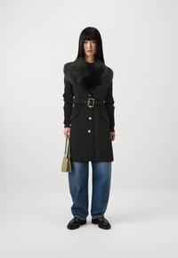 Sort frakke med faux fur krave, bælte i taljen, knapper, sidelommer, parret med løse blå jeans og en lille grøn skuldertaske.