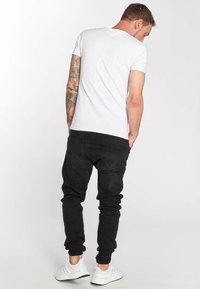 Camiseta blanca de manga corta, jeans negros ajustados y zapatillas blancas. Tatuajes visibles en el brazo izquierdo. Fondo sencillo.