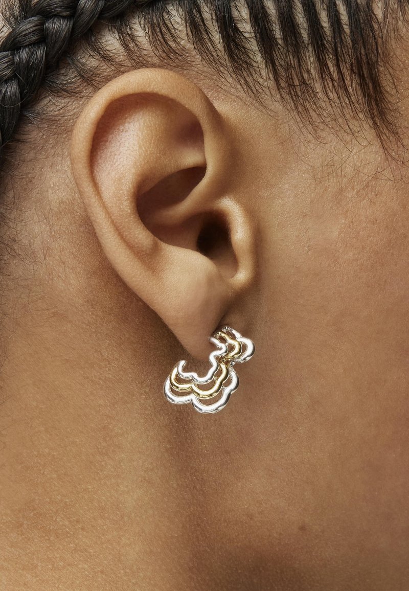 Pendientes ondulados de oro y plata sujetos a una oreja. El diseño presenta formas curvas y en capas con un acabado pulido.