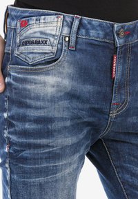 Blaue Denim-Jeans mit einem verblassten Design, ausgestattet mit gesteppten Akzenten, einer markierten Gesäßtasche und roten Hardware-Details.
