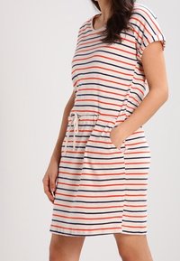 Robe rayée en beige avec des lignes horizontales rouges et navy, à manches courtes, taille élastique avec cordon de serrage et poches latérales. Tissu doux en mélange de coton.