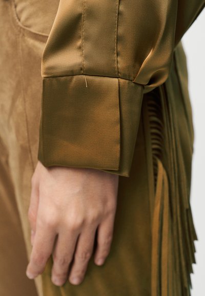 Μανσέτα πουκάμισου από σατέν σε ελιά πράσινο, με κομψή, ομαλή υφή και δομημένο σχέδιο, με εμφανείς ραφές και απαλές λεπτομέρειες με φουντίτσες.