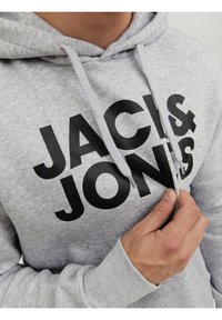 Gros plan d'une personne ajustant le cordon d'un sweat à capuche gris clair avec un grand texte noir « JACK & JONES » sur le devant.