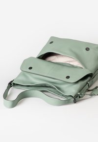 Borsa a tracolla in pelle verde chiaro con il risvolto anteriore aperto, che mostra l'interno rivestito in tessuto beige con motivo, posata su una superficie bianca.