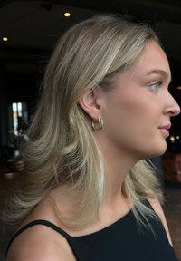 Guldörhänge med en vriden design, visat på en person med långt blont hår, klädd i en svart topp, i en mjukt upplyst inomhusmiljö.