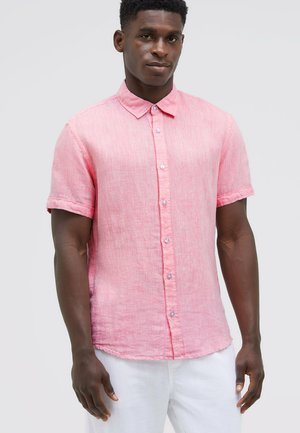 Mann trägt ein rosa Leinenhemd mit kurzen Ärmeln und Knöpfen, mit hochgekrempelten Ärmelbündchen und weißen Hosen vor einem schlichten Hintergrund.