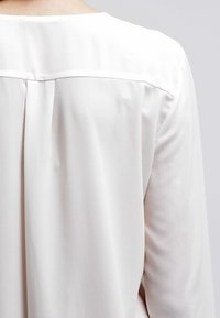 Cremefarvet bluse lavet af et glat, let stof. Har en subtil rygfold og en let glans. Minimalistisk design med en rund halsudskæring.