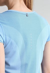 Camiseta deportiva de manga corta en azul claro, hecha de tela de malla, con un acento reflectante cerca del cuello y una fila de pequeños puntos reflectantes.