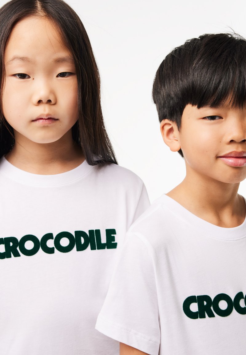Deux enfants portent des t-shirts blancs avec des lettres en relief vert vif qui épellent "CROCODILE". Le tissu semble doux et lisse.