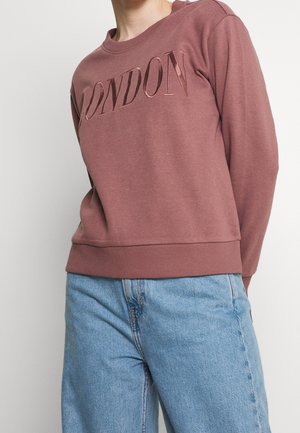 Osoba ubrana w wrzosową bluzę z napisem "LONDON" dużymi literami oraz jasnoniebieskie dżinsy na jednolitym tle.