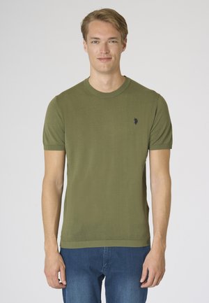 Giovane uomo con capelli biondi corti che indossa un maglione verde oliva a maniche corte e jeans blu su sfondo chiaro.