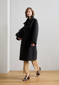 Svart trenchcoat med dobbeltknappede knapper, båret over lysebrune slim bukser og svarte stroppesandaler med en teksturert blokkhel. Bærer en svart håndveske.