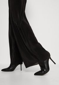Pantalones amplios de pierna plisada negros combinados con botines negros de tacón stiletto en punta sobre un fondo blanco.