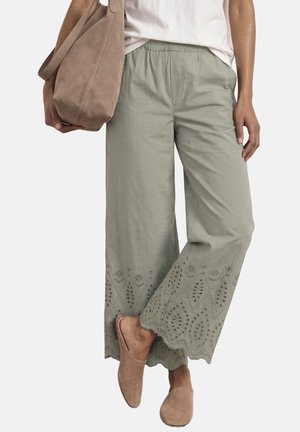 Kvinde iført lysegrå vidde bukser med hulmønsterbroderi, der holder en tan farvet ruskindstaske og har matchende tan slip-on sko på.