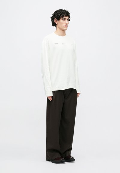 T-shirt de manga comprida creme com texto no peito, combinada com calças largas castanho escuro e sapatos pretos com sola texturizada.