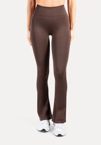 Braune, enganliegende Yogahosen mit hohem Bund, aus glattem Material. Am unteren Ende leicht ausgestellt. Kombiniert mit weißen Turnschuhen.