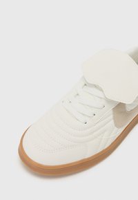 Baskets blanches avec texture matelassée, accents crème et sangle velcro. Comprend des lacets blancs et une semelle en caoutchouc marron clair.