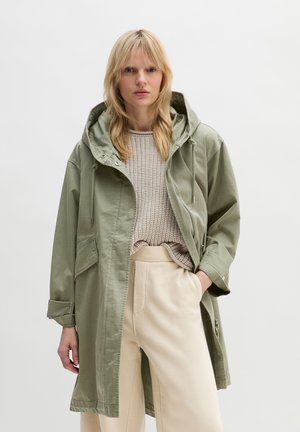 Blonde Frau trägt eine hellgrüne Kapuzenparka, einen beigen Strickpullover und cremefarbene weite Hose, steht vor einem einfarbigen Hintergrund.