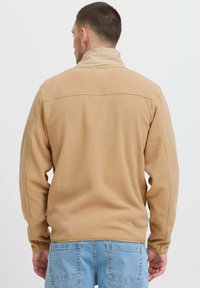 Beige fleece jacket with a high collar, smooth texture, and a seam across the back. Paired with light blue denim pants.