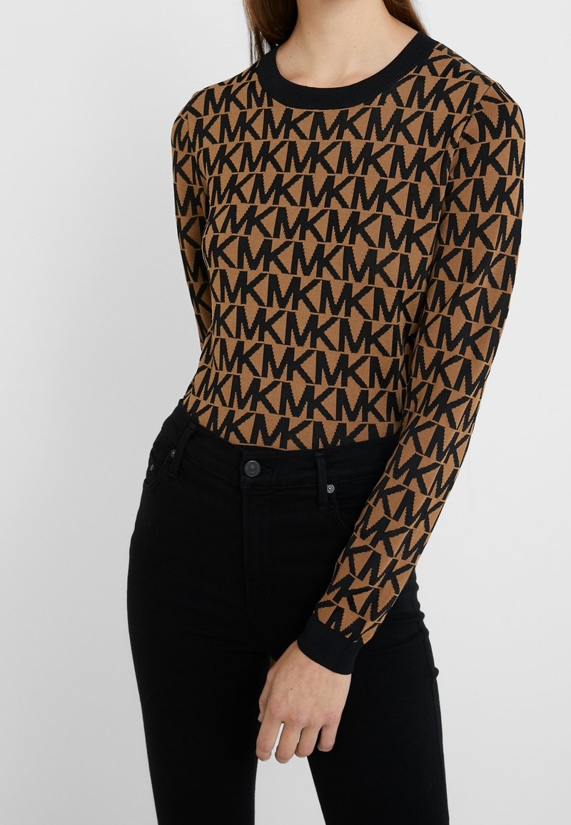 Brauner Strickpullover mit einem sich wiederholenden schwarzen "MK"-Logo-Muster, rundem Ausschnitt und schwarzen Bündchen. Kombiniert mit schwarzen Hosen.