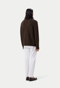 Braun strukturiertes Strickpullover, Rundhalsausschnitt, gerippte Bündchen und Saum, kombiniert mit weißen Hosen und dunklen Slippern, von hinten gesehen.