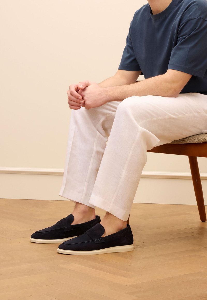 Marineblaue Wildlederschlupfschuhe mit einer beigen Gummisohle, getragen mit hellcremfarbigen Leinenhosen, sitzend auf einem Holzhstuhl vor einem neutralen Hintergrund.