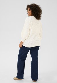 Pull en tricot crème avec un motif en zigzag, manches bouffantes, associé à un jean évasé bleu foncé et des chaussures beiges. Fond clair.