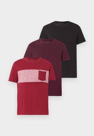 Άνδρας που σκύβει ελαφρώς μπροστά με το ένα χέρι στην τσέπη, φοράει σκούρο μωβ μπλουζάκι, ανοιχτό μπλε τζιν και μαύρα παπούτσια, στέκεται σε λευκό πάτωμα.