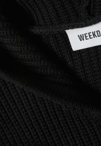 Fekete kötött pulóver texturált mintával és egy fehér "WEEKDAY" feliratú címkével. Az anyag vastagnak és kényelmesnek tűnik.