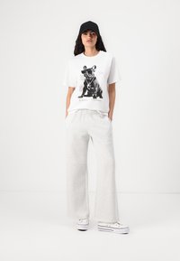 White t-shirt with a black print of a dog, paired with light grey wide-leg sweatpants and white sneakers. Black cap completes the look.