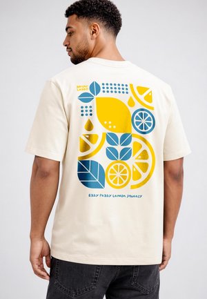 Man wearing a cream t-shirt with colorful lemon, leaf, and drop graphics on the back, text reads "Easy Peasy Lemon Squeezy."