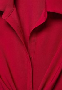 Chemise rouge à col avec une texture lisse, dotée d'une fermeture à boutons et d'une coupe structurée, mettant en valeur les détails de couture sur le col.