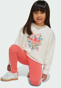Kremowa bluza z kwiatową grafiką; koralowe legginsy; szaro-białe trampki z gumową podeszwą; pozycja klęcząca; miękkie tekstury, casualowy design.